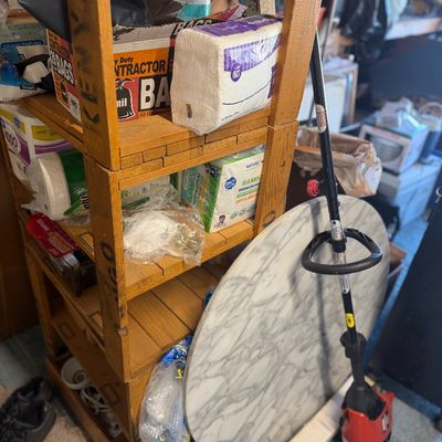 Sale Photo Thumbnail #202: Rustic wooden shelving unit stocked with paper products, standing beside a round marble-look tabletop and a gas string trimmer.