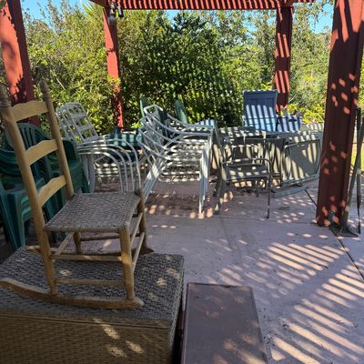 Sale Photo Thumbnail #67: Collection of outdoor patio furniture under a wooden pergola, featuring a child's woven-seat rocking chair, stacked metal lawn chairs, plastic patio chairs, and storage trunks.