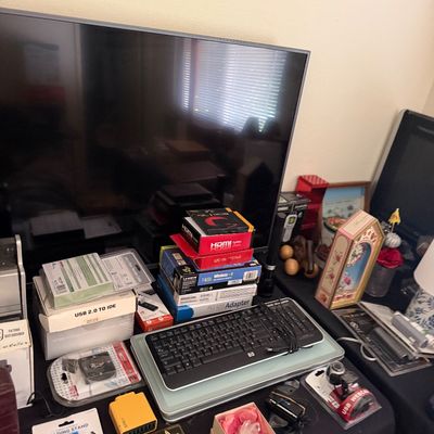 Sale Photo Thumbnail #194: Collection of assorted electronics and computer accessories, including an HP keyboard, adapters, cables, and small boxed gadgets arranged on a table in front of a flat-screen TV.