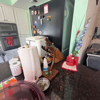 Sale Photo Thumbnail #89: Kitchen countertop with various household goods, including a wooden knife block, paper towel holder, red vase with faux flowers, and a black refrigerator in the background.