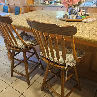 Sale Photo Thumbnail #40: Pair of vintage oak swivel bar stools featuring spindle backs and floral tie-on cushions, positioned at a granite kitchen island.