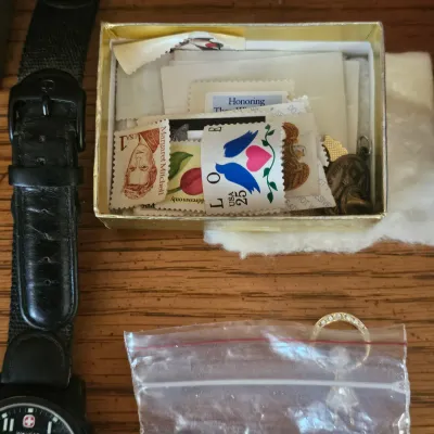 Sale Photo Thumbnail #49: Collection of small trinkets, including vintage US postage stamps, a Wenger watch strap, and a small plastic bag with jewelry, resting on a wooden surface.