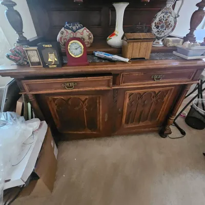 Sale Photo Thumbnail #97: Vintage dark wood buffet server featuring gothic arched panel doors, brass hardware, and a collection of tabletop decor including clocks, vases, and a pitcher.