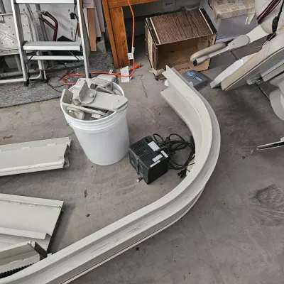 Sale Photo Thumbnail #55: Disassembled motorized stair lift components, including curved track sections, mounting brackets in a bucket, and power supply unit.