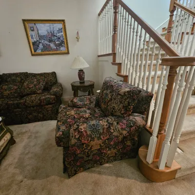 Sale Photo Thumbnail #9: Traditional floral tapestry upholstered armchair and matching sofa set, positioned near an entryway staircase with a wood end table and table lamp.