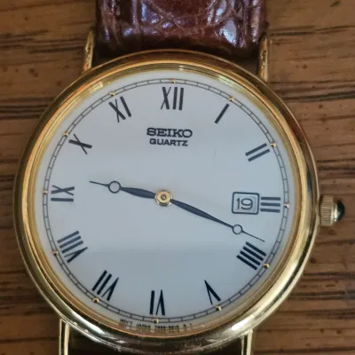 Sale Photo Thumbnail #48: Vintage Seiko Quartz dress watch featuring a gold-tone round bezel, white dial with Roman numeral hour markers, date window at 3 o'clock, and a brown leather strap.