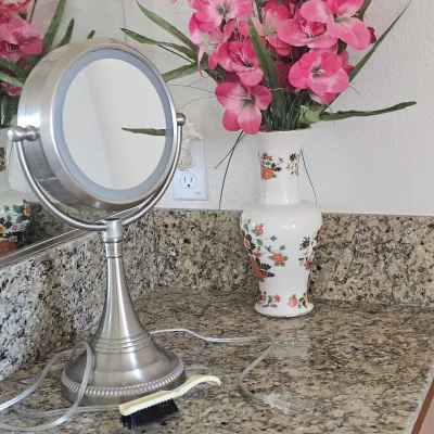 Sale Photo Thumbnail #63: Brushed nickel tabletop illuminated vanity mirror with power cord, shown alongside a small cleaning brush and a floral porcelain vase.