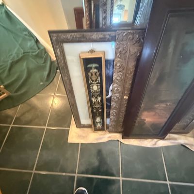 Sale Photo Thumbnail #160: Collection of framed artwork and ornate mirrors leaning against a wall, including a narrow vertical panel with intricate classical motifs.