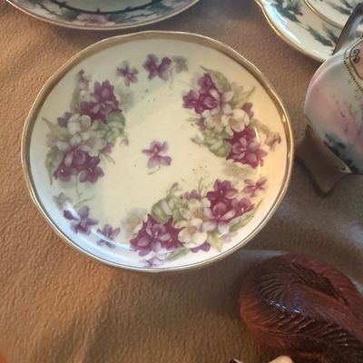 Sale Photo Thumbnail #112: Vintage porcelain saucer featuring hand-painted purple floral designs and a gilded rim, displayed alongside other decorative tableware.
