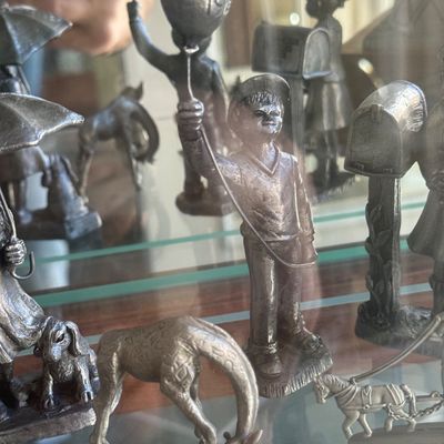 Sale Photo Thumbnail #145: Collection of pewter metal figurines, featuring a young boy holding a balloon, a giraffe, children with umbrellas, and a horse-drawn carriage displayed on glass display case shelves.