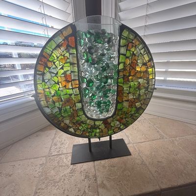 Sale Photo Thumbnail #10: Circular mosaic stained glass decorative piece mounted on a metal stand, featuring green and amber tiles surrounding a central glass cylinder filled with decorative glass beads.