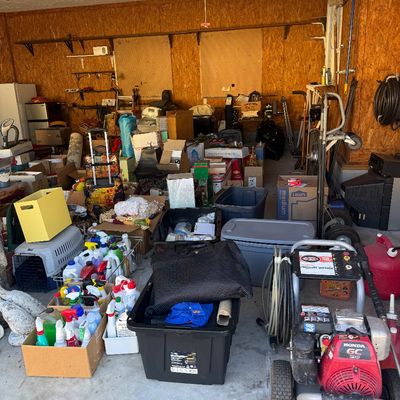 Sale Photo Thumbnail #13: A garage filled with estate sale items including a Simpson gas-powered pressure washer, plastic storage totes, household cleaning supplies, a hand truck, and various boxed goods.