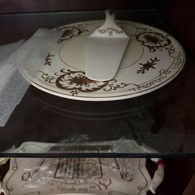 Sale Photo Thumbnail #38: Vintage ceramic cake plate and matching server set featuring a dark gold floral scrolled motif, displayed on a glass shelf above another wrapped serving tray.