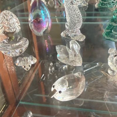 Sale Photo Thumbnail #137: Collection of lead crystal and art glass figurines, including Swarovski-style animals, a seahorse, egg paperweights, and a green glass tree, displayed on a glass shelf.