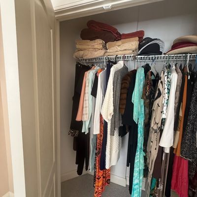 Sale Photo Thumbnail #163: A reach-in closet filled with assorted women's clothing, including dresses, sweaters, and jackets, alongside folded linens and hats on the upper shelf.