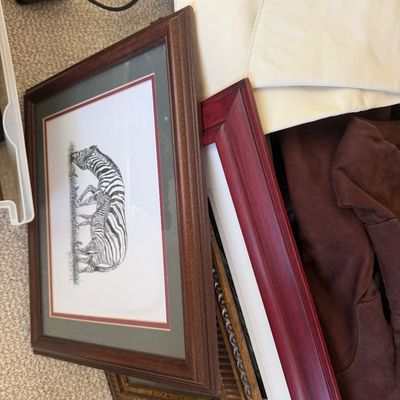 Sale Photo Thumbnail #25: Collection of framed artwork, including a matted zebra sketch print in a wood frame, stacked alongside other picture frames and clothing items.