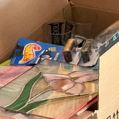 Sale Photo Thumbnail #18: Cardboard box containing a stained glass flower suncatcher, packaged Hot Wheels die-cast cars, and printed glassware.