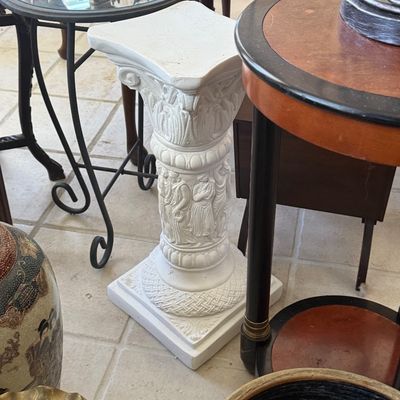 Sale Photo Thumbnail #74: White Classical-style plaster column pedestal with relief figures along the shaft and a square top, surrounded by small side tables.