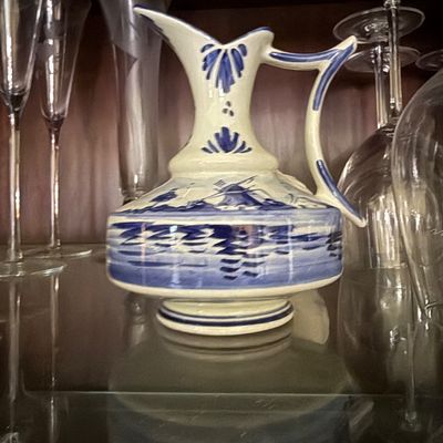 Sale Photo Thumbnail #35: Hand-painted Delftware style ceramic pitcher with windmill graphics, displayed on a glass shelf alongside crystal stemware.
