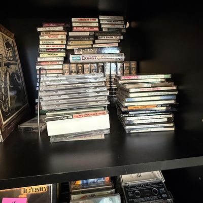 Sale Photo Thumbnail #79: Collection of music CDs and cassette tapes arranged on black shelving units, featuring various artists from jazz, pop, and country genres.