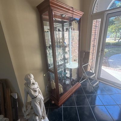 Sale Photo Thumbnail #146: Tall traditional wood-frame glass curio cabinet filled with various collectibles, positioned beside a classical-style female statue sculpture in an entryway.