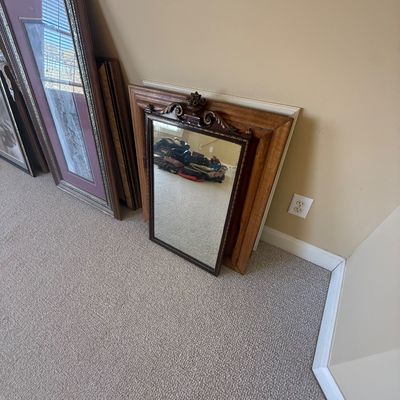 Sale Photo Thumbnail #169: Collection of framed artwork and mirrors leaning against a wall, including a small ornate wall mirror with carved crest detail and a large silver-toned framed picture.