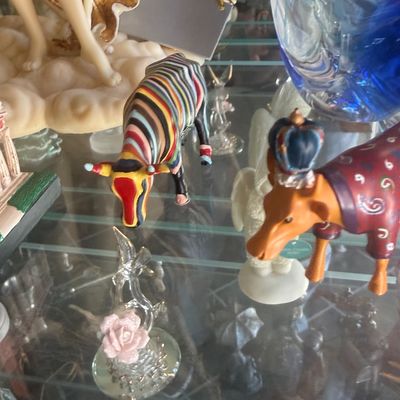 Sale Photo Thumbnail #129: Collection of decorative figurines displayed on glass shelves, including two colorful CowParade cows, a blown glass rose, and a large blue glass vase.
