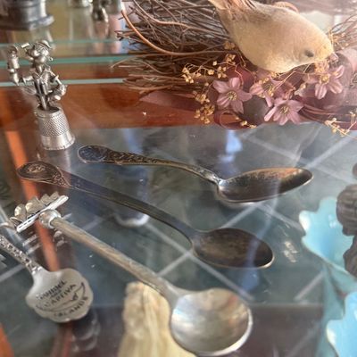 Sale Photo Thumbnail #141: Collection of souvenir demitasse spoons, silver-plated knick-knacks, a decorative bird's nest, and a novelty character thimble displayed in a glass cabinet.