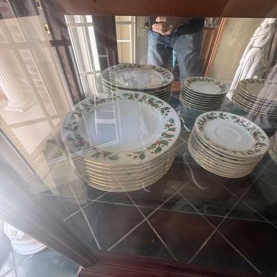 Sale Photo Thumbnail #148: Holiday holly motif porcelain china service set displayed inside a wooden cabinet, featuring stacked dinner plates and saucers with gold trim.