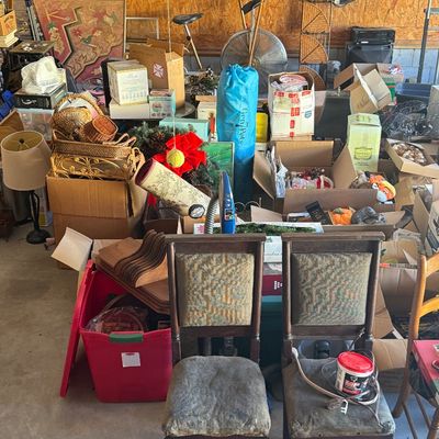 Sale Photo Thumbnail #15: A garage packed with household goods, including a pair of antique upholstered dining chairs in the foreground, various cardboard boxes, lamps, and a vintage exercise bike.