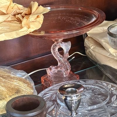 Sale Photo Thumbnail #57: Vintage pink depression glass cake stand featuring a carved koi fish pedestal base, displayed alongside clear cut glass plates and a silver candlestick.