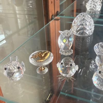 Sale Photo Thumbnail #138: Collection of crystal miniature figurines displayed on glass shelves, including birds, an owl, and a small fruit bowl with gold-tone accents.