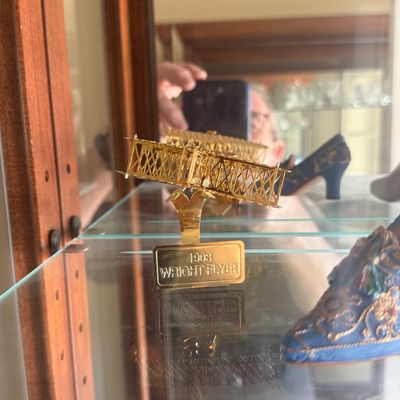 Sale Photo Thumbnail #120: Miniature brass 1903 Wright Flyer model on a display stand, presented in a glass cabinet alongside decorative collectible shoes.