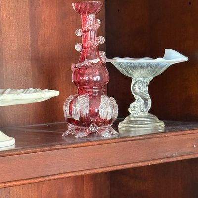 Sale Photo Thumbnail #58: Collection of vintage art glass, featuring a cranberry glass vase with clear applied rigaree and leaf details beside an opalescent compote with a dolphin-shaped pedestal.
