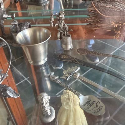Sale Photo Thumbnail #142: Collection of vintage pewter and metal miniatures displayed on a glass shelf, including souvenir spoons, a thimble topped with a sailor figure, small cups, and decorative figurines.