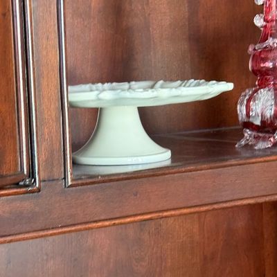 Sale Photo Thumbnail #59: Vintage white milk glass pedestal cake stand displayed inside a wooden cabinet alongside an ornate ruby glass vase.