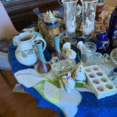 Sale Photo Thumbnail #41: A collection of vintage home decor items displayed on a floral tablecloth, including ceramic vases, beer steins, glass pilsner glasses, animal figurines, and snow globes.
