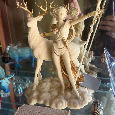 Sale Photo Thumbnail #131: Ivory-toned resin figurine of Diana the Huntress with a stag, featuring gilt accents and a cloudy base.