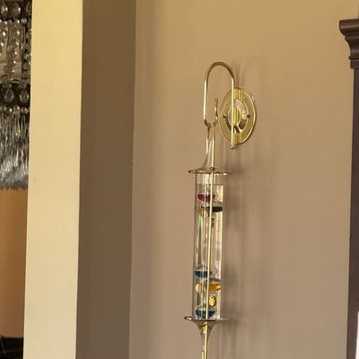 Sale Photo Thumbnail #77: Wall-mounted glass Galileo thermometer suspended from a polished brass hook, featuring colorful floating spheres.