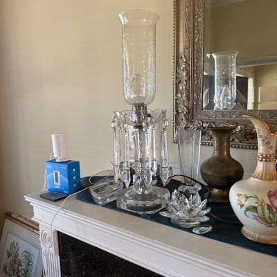 Sale Photo Thumbnail #36: Vintage crystal hurricane table lamp with hanging prism drops, displayed on a mantelpiece alongside various glassware, a floral vase, and a brass vessel.