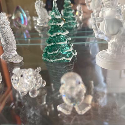Sale Photo Thumbnail #134: Collection of glass and crystal figurines, including green glass snow-capped Christmas trees, crystal animals, and a frosted glass child figurine on glass display shelves.