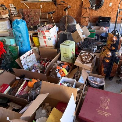 Sale Photo Thumbnail #95: A garage sale setup packed with numerous cardboard boxes filled with household goods, seasonal decor, stuffed animals, glassware, luggage, and vintage items.