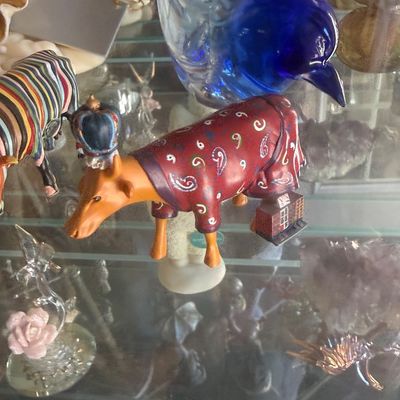 Sale Photo Thumbnail #130: CowParade painted cow figurine wearing a crown and a paisley jacket, displayed on a glass shelf alongside a cobalt blue glass bird.
