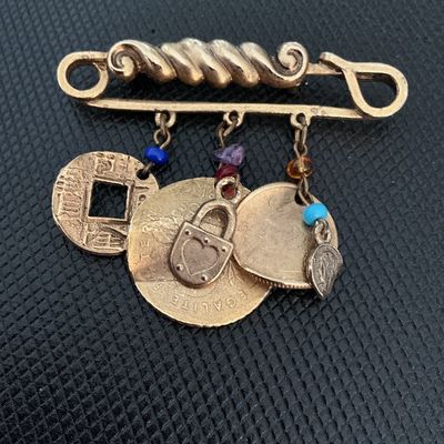 Sale Photo Thumbnail #161: Gold-tone safety pin style brooch featuring several dangling charm components, including coin replicas (such as a Chinese square-hole coin and French coin style pieces), a heart padlock charm, a small medallion, and colorful accent beads. Shows light wear