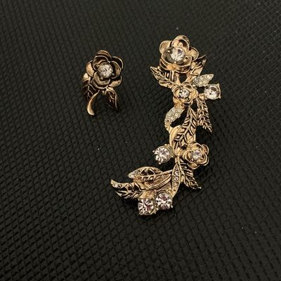 Sale Photo Thumbnail #178: An asymmetrical fashion jewelry earring set featuring a single stud earring and an elongated floral ear crawler design. Crafted in gold-tone metal and accented with clear rhinestones in flower and leaf motifs. Pierced post backs (earring backs/nuts not in