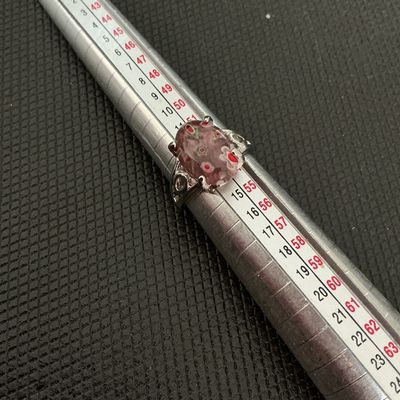 Sale Photo Thumbnail #204: A silver-tone ring featuring a bezel/prong-set oval millefiori glass cabochon with red, green, and white floral design elements inside a pinkish glass base. Shows minor surface wear consistent with age. Size measures approximately 6.5 - 6.75 on a ring man