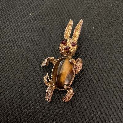 Sale Photo Thumbnail #226: Vintage gold-tone textured metal brooch featuring a playful bunny rabbit design. The body is set with an oval tiger's eye cabochon, and the head features red rhinestone eyes and nose details. Standard pin back closure with pin mechanism. Shows light vinta