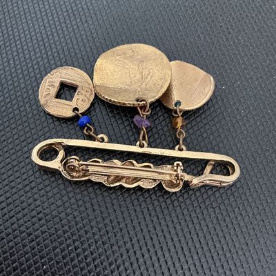 Sale Photo Thumbnail #162: Gold-tone safety pin style brooch featuring several dangling charm components, including coin replicas (such as a Chinese square-hole coin and French coin style pieces), a heart padlock charm, a small medallion, and colorful accent beads. Shows light wear