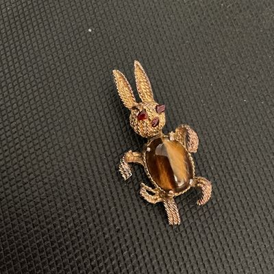Sale Photo Thumbnail #227: Vintage gold-tone textured metal brooch featuring a playful bunny rabbit design. The body is set with an oval tiger's eye cabochon, and the head features red rhinestone eyes and nose details. Standard pin back closure with pin mechanism. Shows light vinta