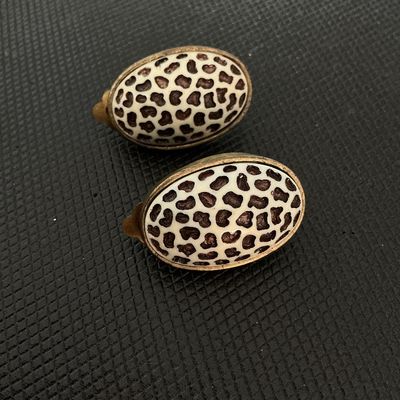 Sale Photo Thumbnail #220: Pair of vintage oval-shaped clip-on earrings featuring a cream and dark brown leopard/cheetah animal print pattern in enamel or resin with a antique brass-tone metal backing and clip hardware. Shows light age-appropriate wear and patina on the metal hardw